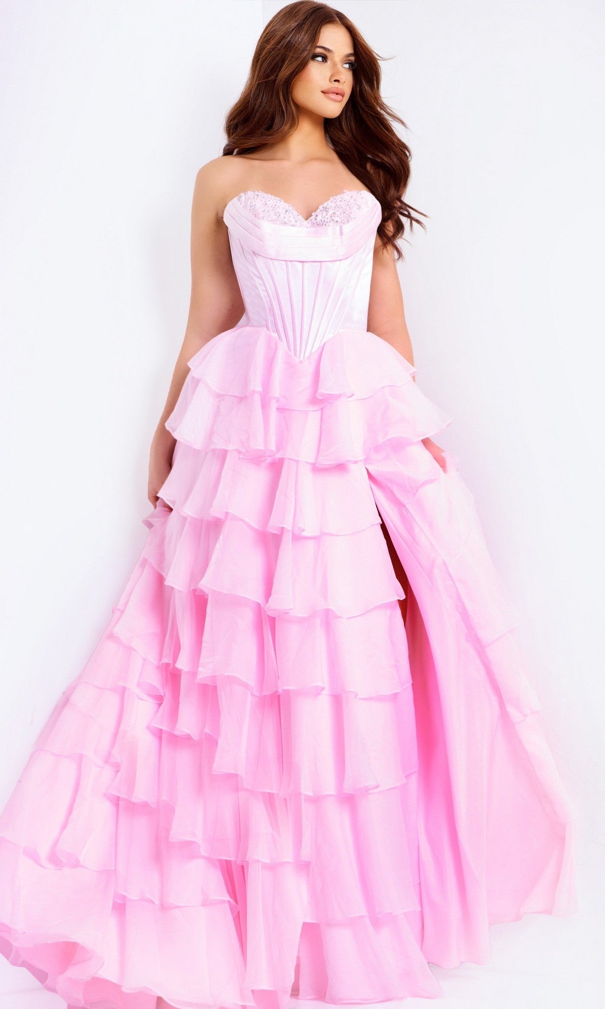 A woman wears the Jovani 47762 Long Prom Dress in pink—strapless, with a satin corset bodice and a ruffle tiered chiffon skirt featuring a high slit—standing against a plain white background.